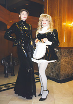 Mistress Amanda Wildefyre in PVC gown and feminized male maid on leash in pvc maid uniform at event