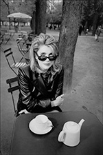an elegant woman peers over her sunglasses, dressed in a fabulous leather coat and enjoying tea in a park